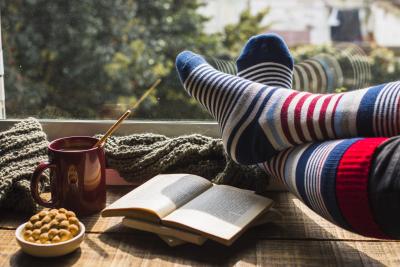photo of person in long socks with book, mug, and snacks looking out window