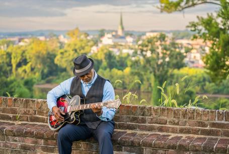 older man playing guitar on a bench with scenic landscape in background