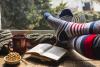 photo of person in long socks with book, mug, and snacks looking out window